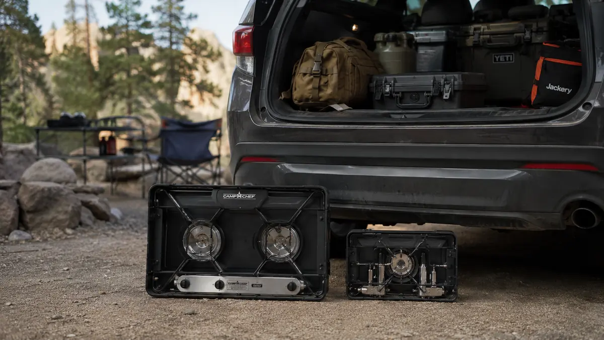 Two folded camp stoves next to the open cargo area of a 4Runner, one small and compact, one noticeably larger