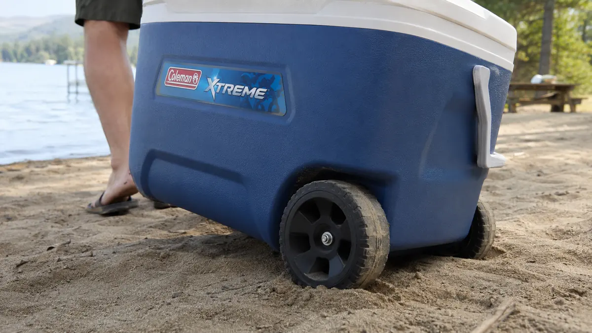 Close-up of the Coleman Xtreme cooler's wheels partially sunk into sand near a lake shore
