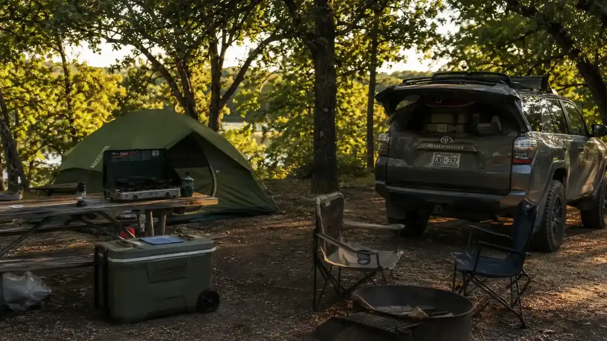 A family campsite at golden hour with a dome tent, camp chairs, and a cooler beside a picnic table under oak trees