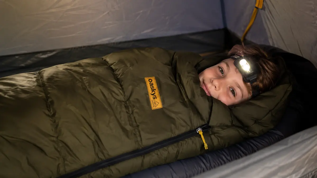 A dark green mummy-style sleeping bag unrolled on a sleeping pad inside a tent, zipper pulled up near the chin