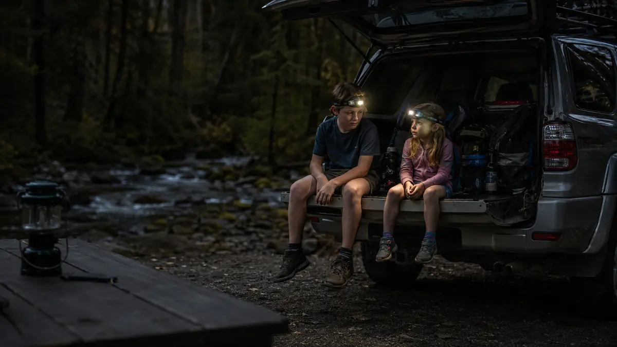 A family camp scene at night with two kids using headlamps instead of a lantern, an unlit lantern sitting dark in the foreground